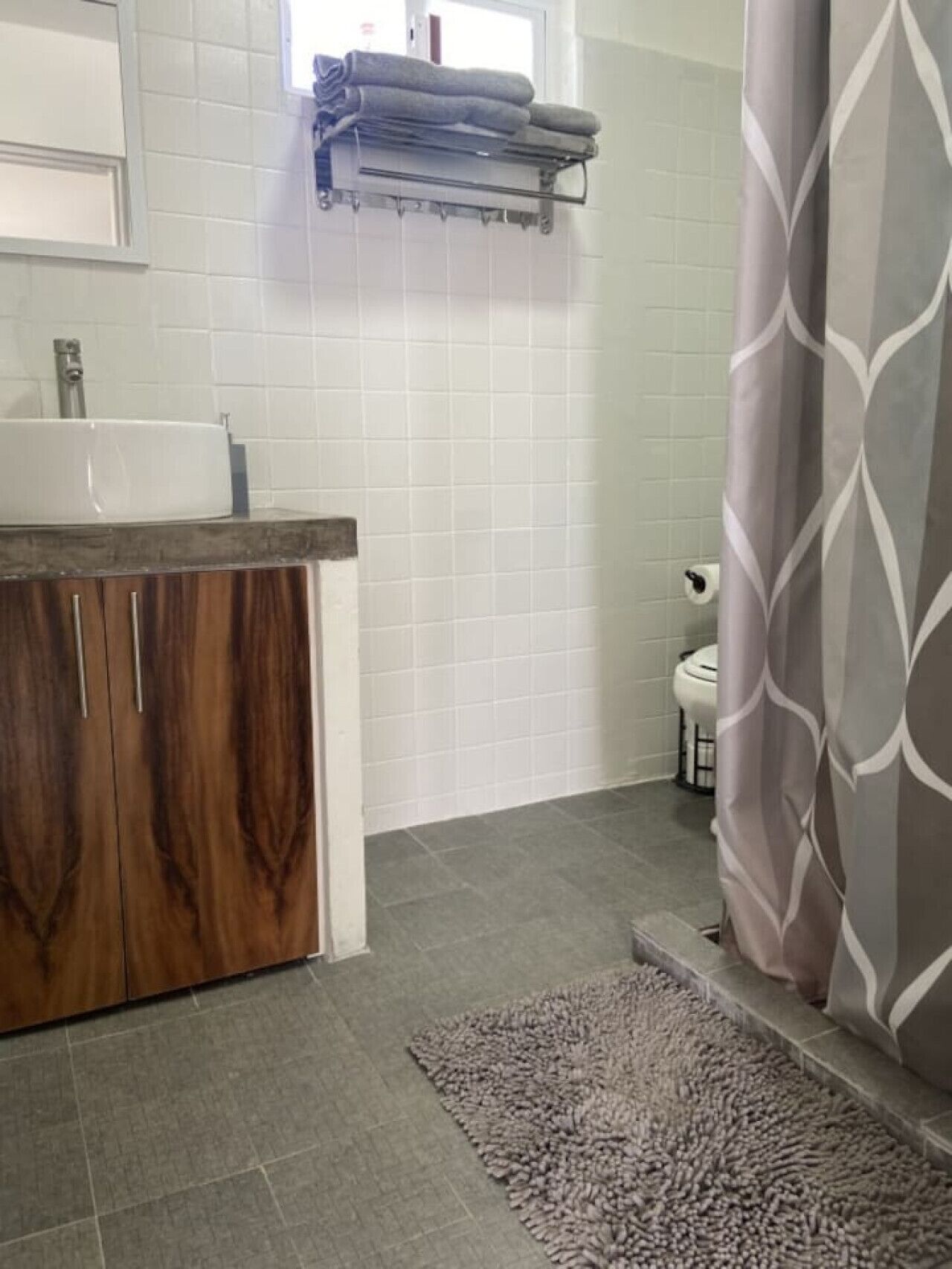 A bathroom with a wooden vanity, white tiles, a towel rack, a toilet, and a shower curtain.