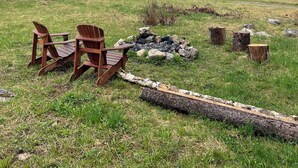 A serene outdoor setting with a wooden table and chairs, a log fire, and a backdrop of snow-capped mountains and dense forest. - Beaverfoot Lodge (Golden)