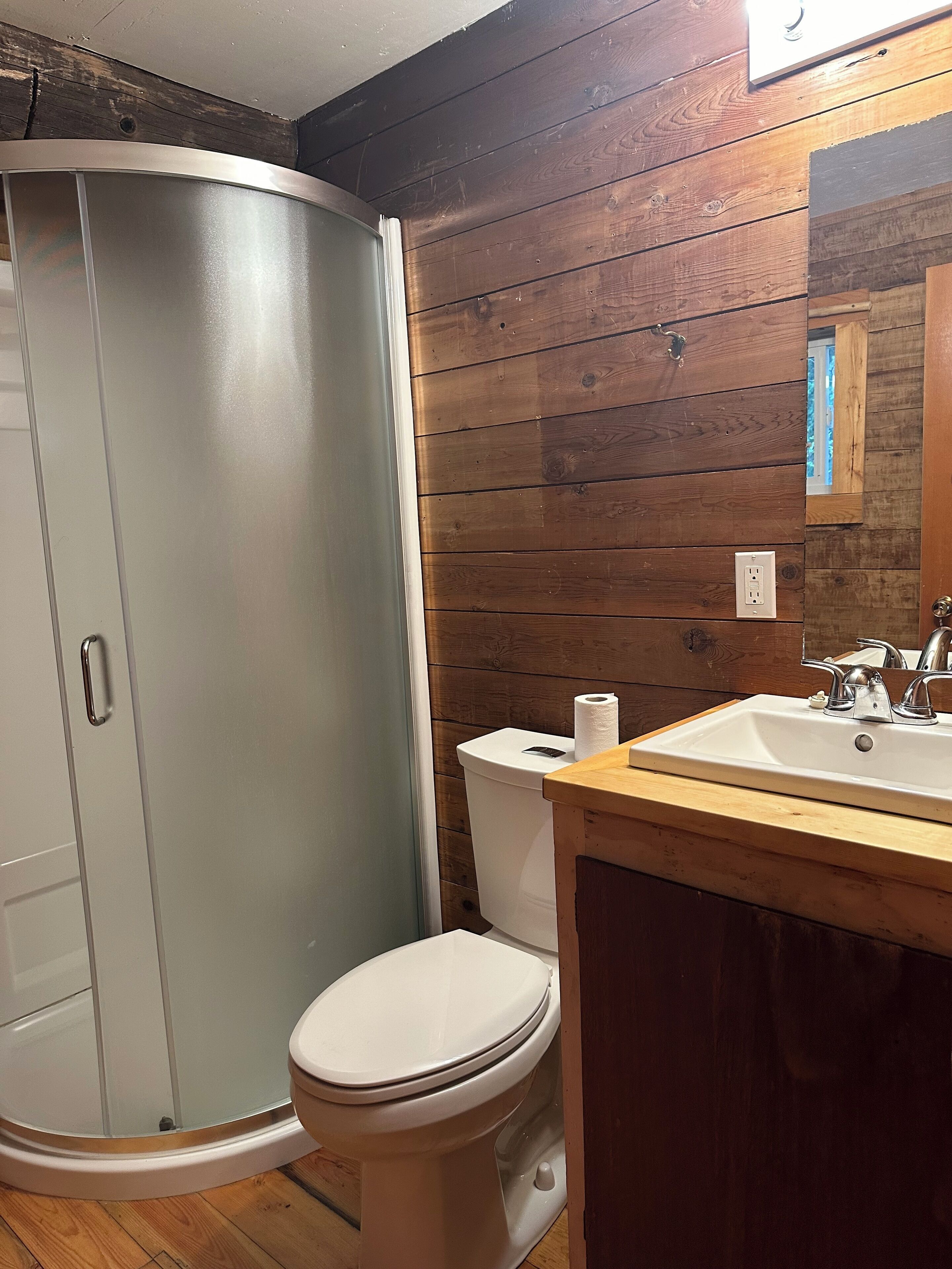 Et badeværelse med glasvæg omkring bruseren, toilet og vask med træbordplade.