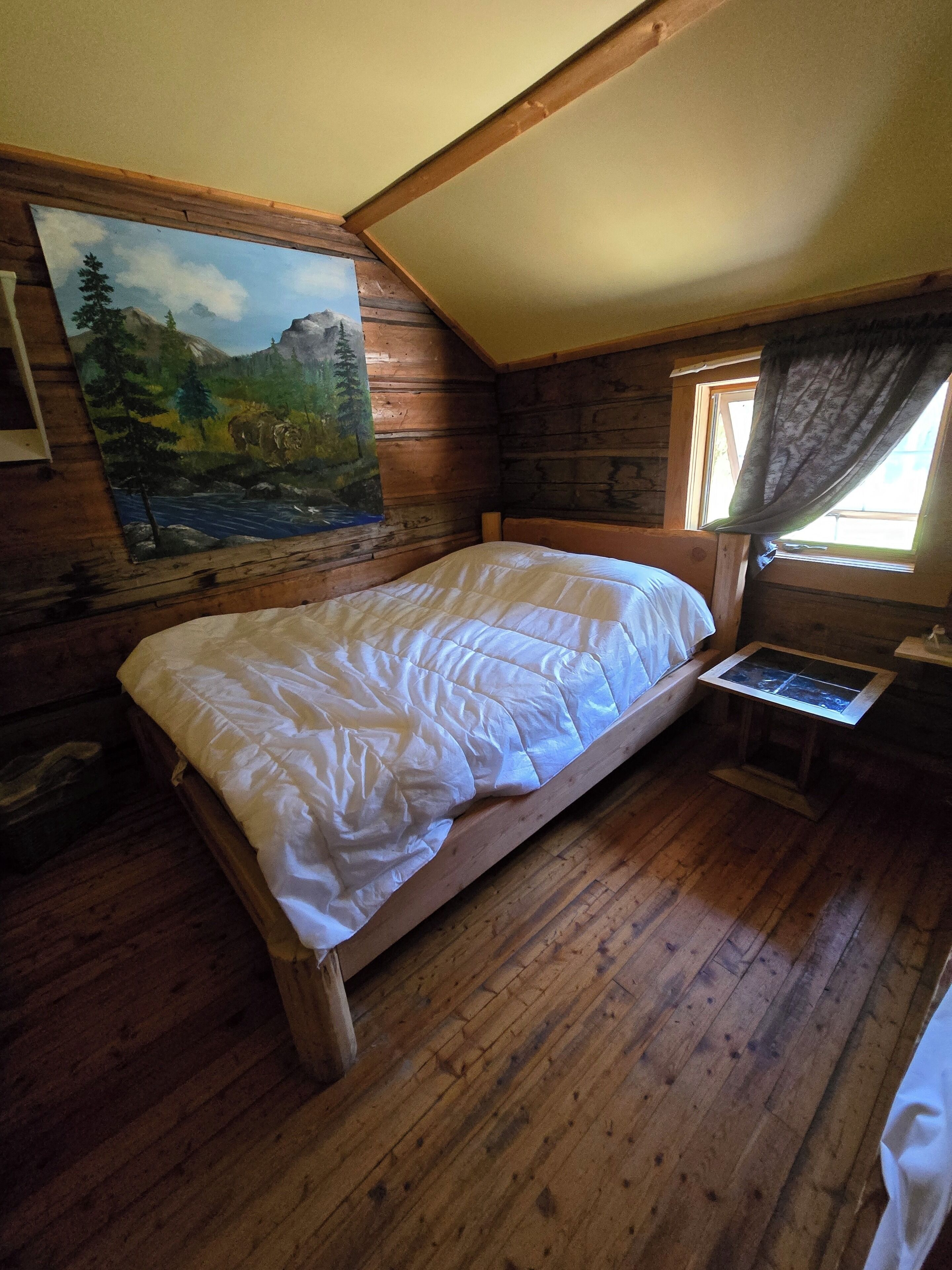 Une chambre à coucher dans une cabane en bois, avec un lit, un tableau représentant un paysage de montagne et une fenêtre avec des rideaux.