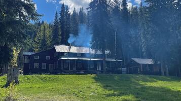 Una cabaña rústica con chimenea humeante, rodeada de un bosque frondoso y un cielo despejado.