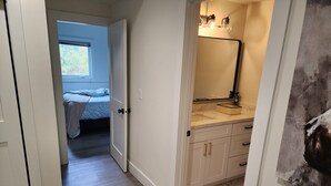 Ein schmaler Korridor mit Holzboden führt zu einem Schlafzimmer mit Bett und Fenster. Rechts ist ein Badezimmer mit Spiegel und Waschbecken zu sehen.