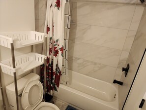 A bathroom with a toilet, a shower curtain with a floral pattern, a bathtub, and a white shelf unit with multiple compartments.
