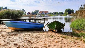 A sandy beach with two boats, a dock, and a calm lake with a house and trees in the background.
