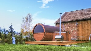 A sauna with a large wooden barrel, a wooden deck, and a brick building in the background.