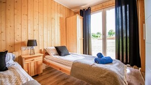 A room with wooden walls and floors, a bed with a white and beige patterned bedspread, a small bedside table with a lamp, and a window with curtains.