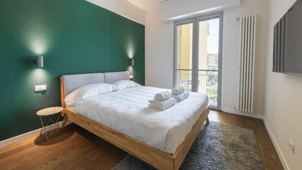 A bedroom with a wooden bed, a small round table, a green wall, a window with a view, and a radiator. - Guido Monaco in Florence by Mmega (Firenze)
