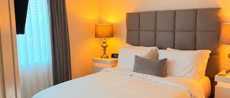 A hotel room with a bed, bedside lamps, a clock, a ceiling fan, and a view of the outside through a window with curtains.