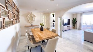 Un salon moderne avec une table à manger en bois, des chaises blanches, un téléviseur et une grande fenêtre avec des rideaux.