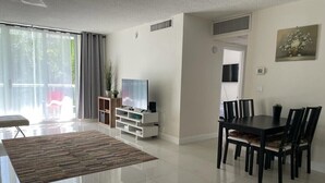 Un salon moderne avec un téléviseur à écran plat, une table à manger et des chaises. Il y a une grande fenêtre avec des rideaux, une plante et un tableau au mur.