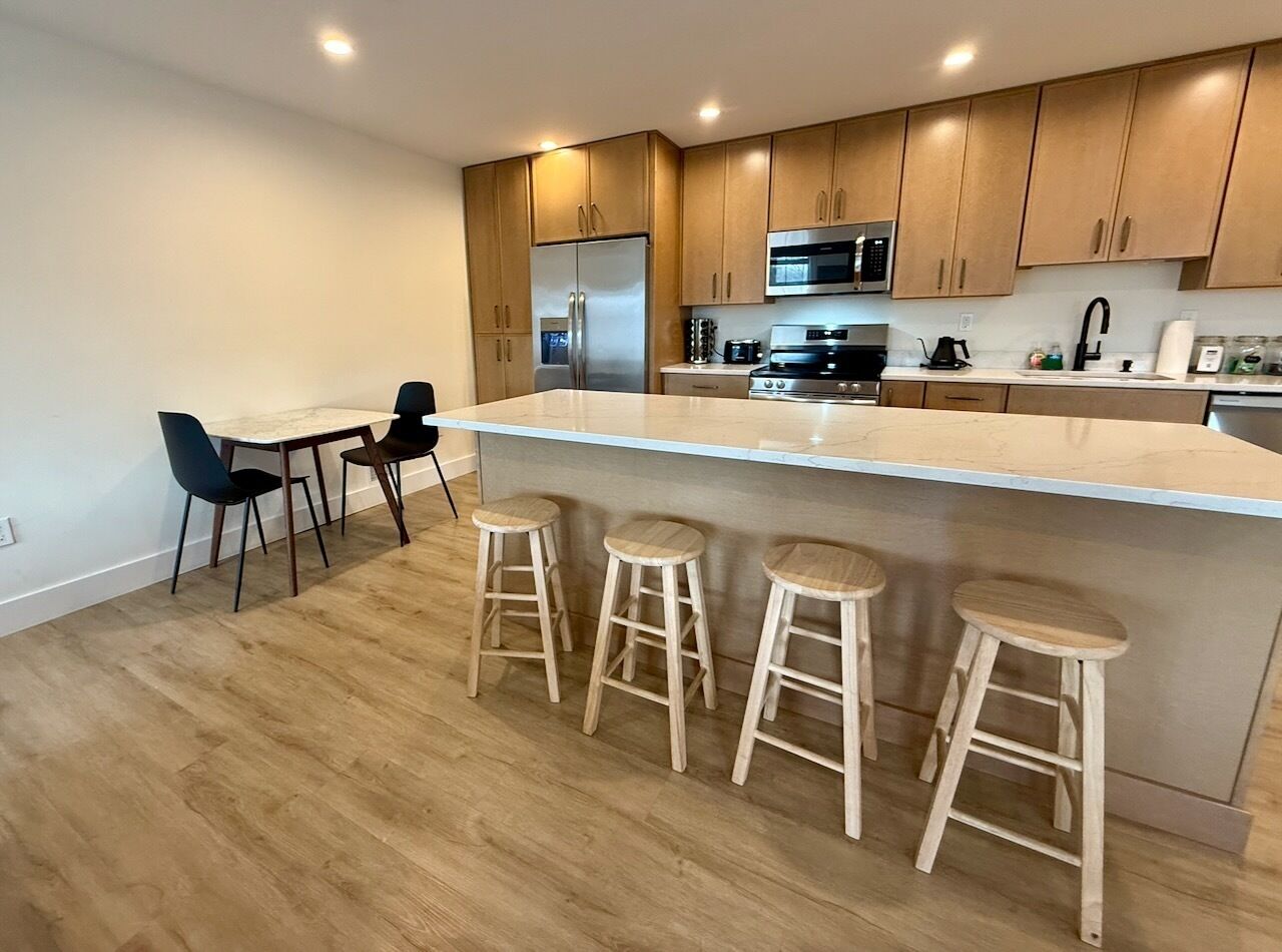 Una cocina moderna con armarios de madera, un refrigerador de acero inoxidable, un microondas y una estufa a gas. Hay tres taburetes de madera y una mesa de comedor con dos sillas.