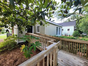 A wooden deck leading to a house with a porch, surrounded by greenery and a fence.