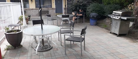 Une terrasse extérieure avec une table ronde et des chaises, un barbecue et une poubelle.