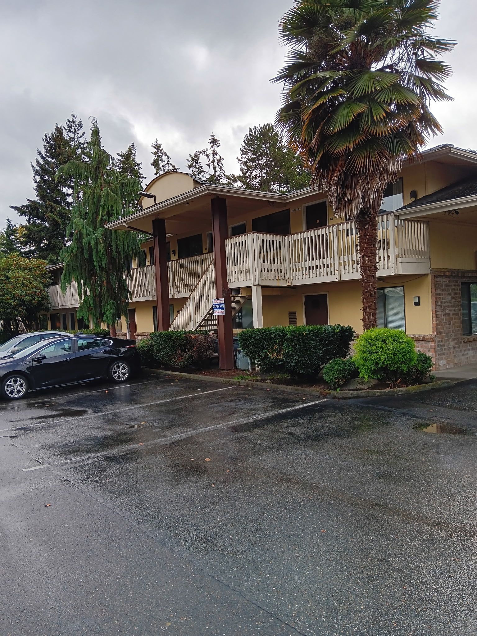 A two-story building with a balcony, a palm tree, and a black car parked in front.