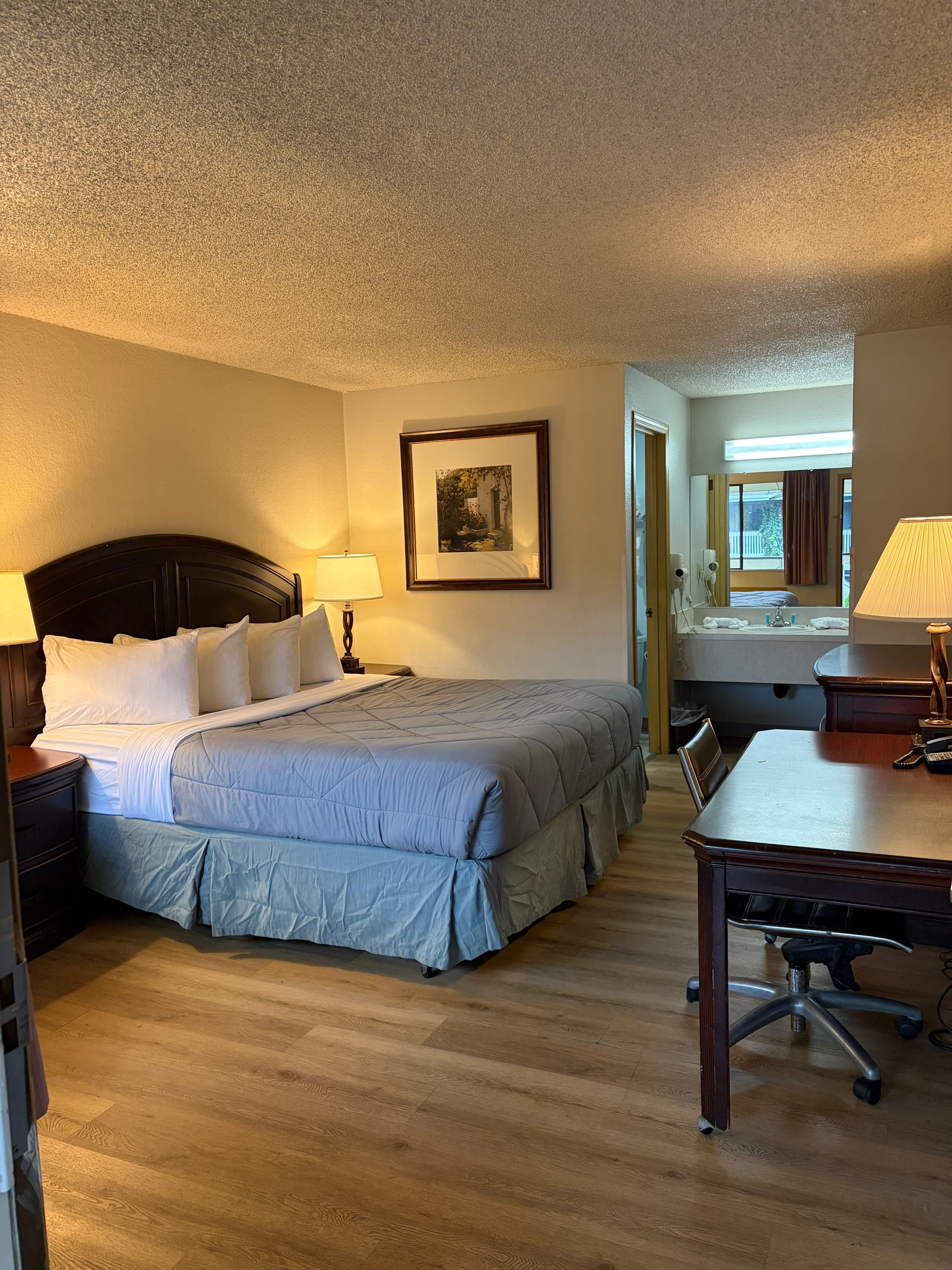 A hotel room with a bed, bedside table, lamp, framed picture, and a desk with a chair.