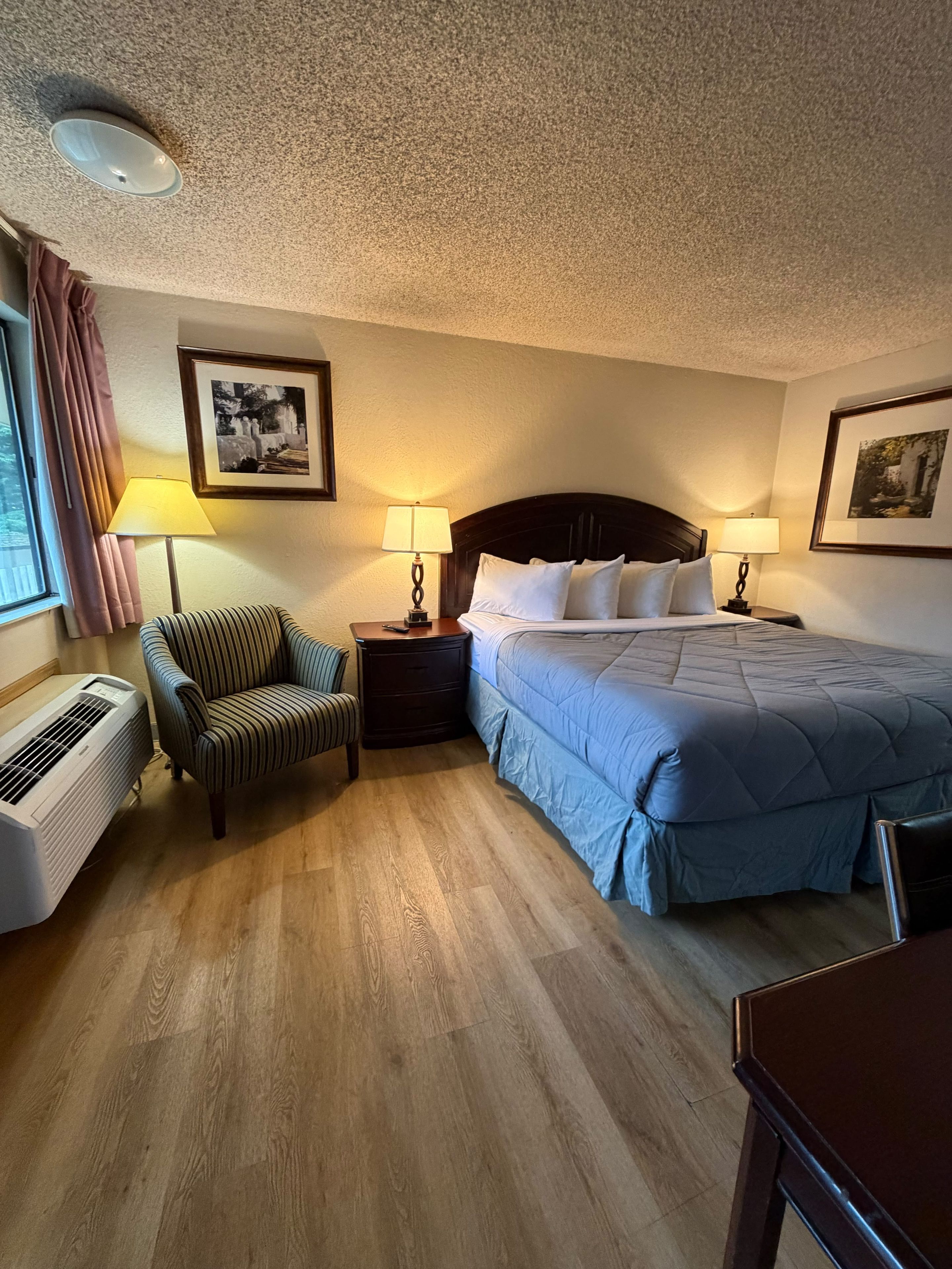 A hotel room with a bed, two bedside tables with lamps, a chair, a heater, and two framed pictures on the walls.