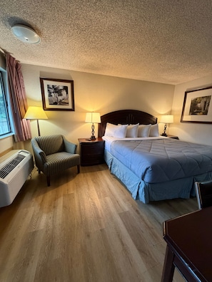 Habitación de hotel con una cama, dos mesitas de noche con lámparas, una silla, un calentador y dos cuadros en las paredes.