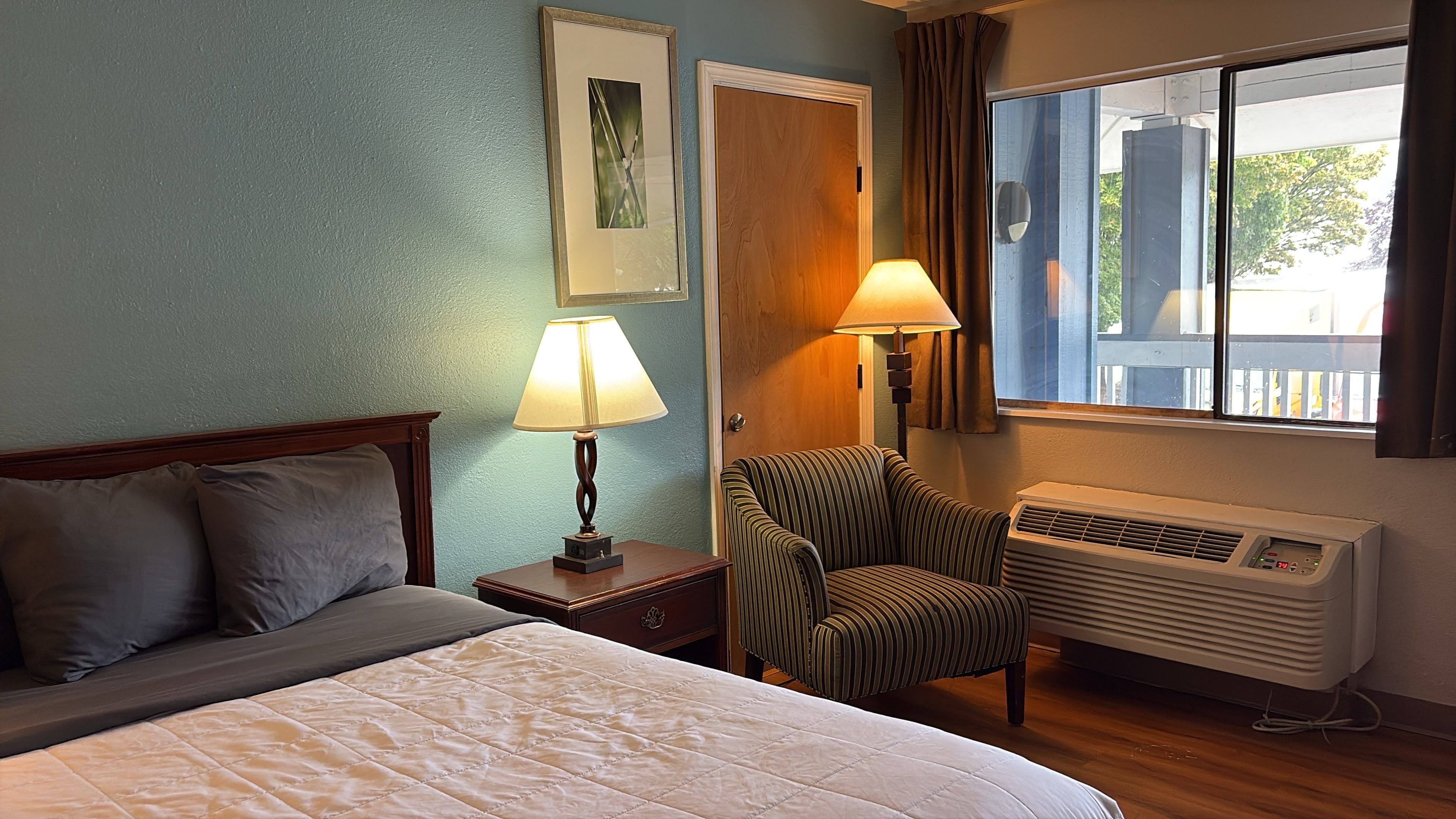 A hotel room with a bed, a chair, a nightstand with a lamp, a wall-mounted air conditioner, and a window with curtains.