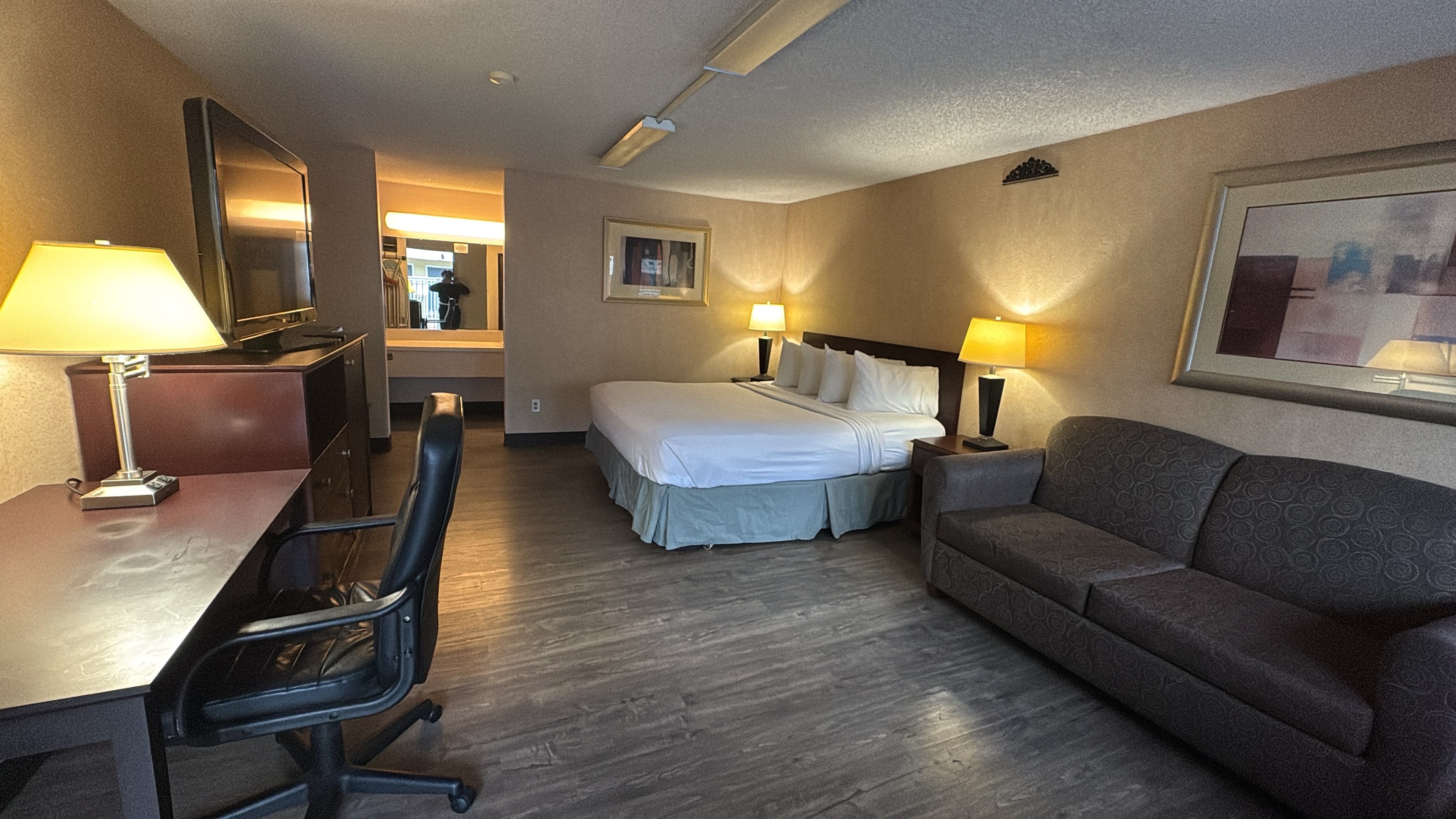 A hotel room with a bed, a desk, a chair, a sofa, and two bedside lamps.