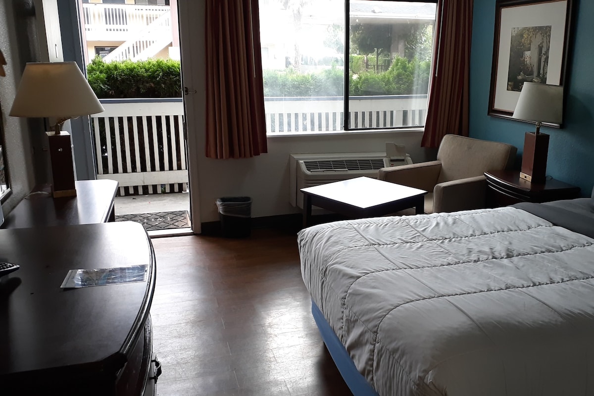 A hotel room with a bed, a desk, a chair, an air conditioning unit, and a window with curtains.