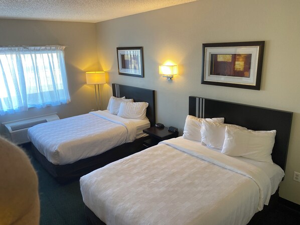 A hotel room with two beds, a window with sheer curtains, a standing lamp, and framed pictures on the wall.