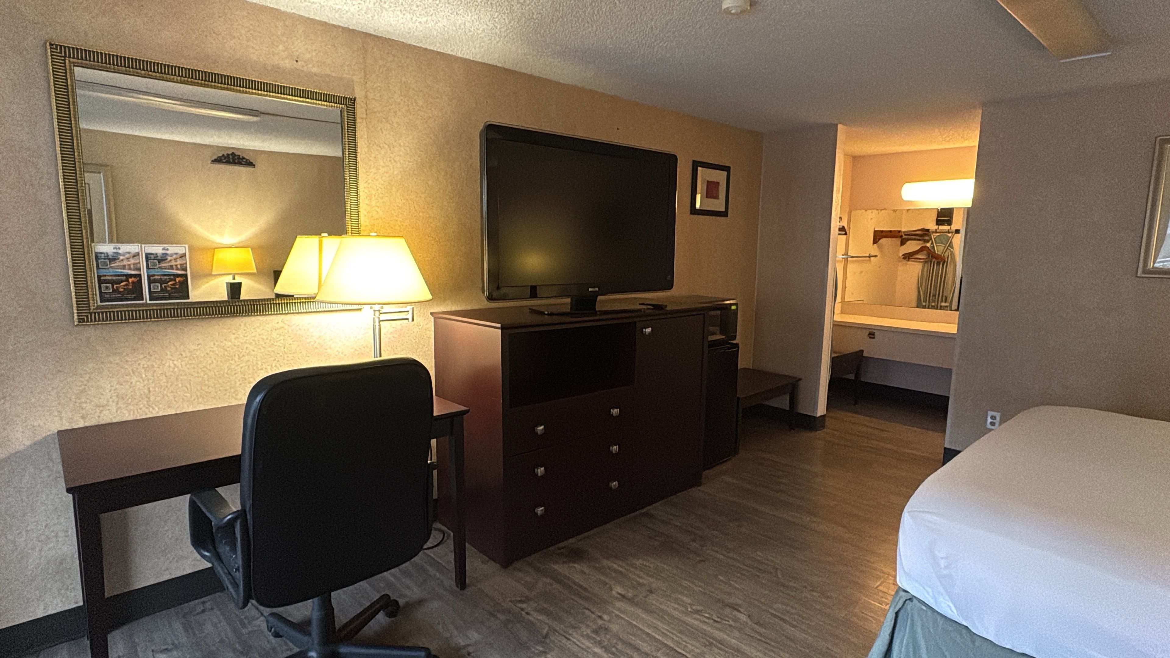Quarto de hotel com cama, escrivaninha, cadeira, televisão, cômoda, espelho e abajur.