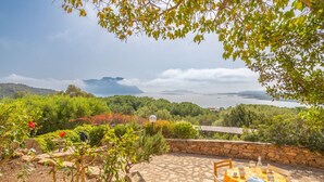 Une terrasse avec un sol en pierre et une table dressée pour les repas, offrant une vue panoramique sur un paysage vallonné et un plan d’eau.