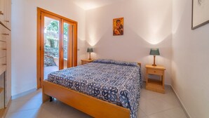 Une chambre à coucher comprenant un lit, deux tables de chevet, une porte donnant sur un espace extérieur et un mur orné d’un tableau encadré.