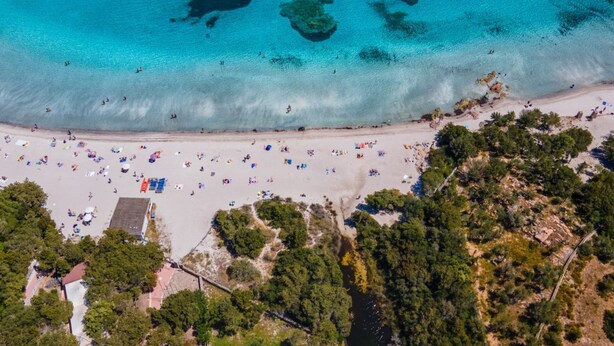 Vue aérienne d’une plage aux eaux turquoise et limpides, bordée d’un rivage sablonneux et parsemée de parasols.