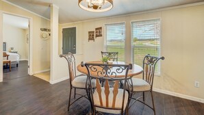 A dining area with a round table, four chairs, and a chandelier. There are two windows with blinds, a door, and a hallway leading to a bedroom. - Hawkeye (Peyton)