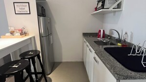 Eine kleine Küche mit Kühlschrank, Spüle und Arbeitsplatte. Zwei Hocker und ein Regal befinden sich über der Spüle.