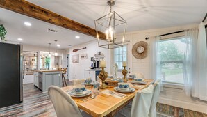 Un comedor con una mesa de madera dispuesta para cuatro personas, rodeada de sillas blancas. Se ve una cocina con isla integrada al fondo.