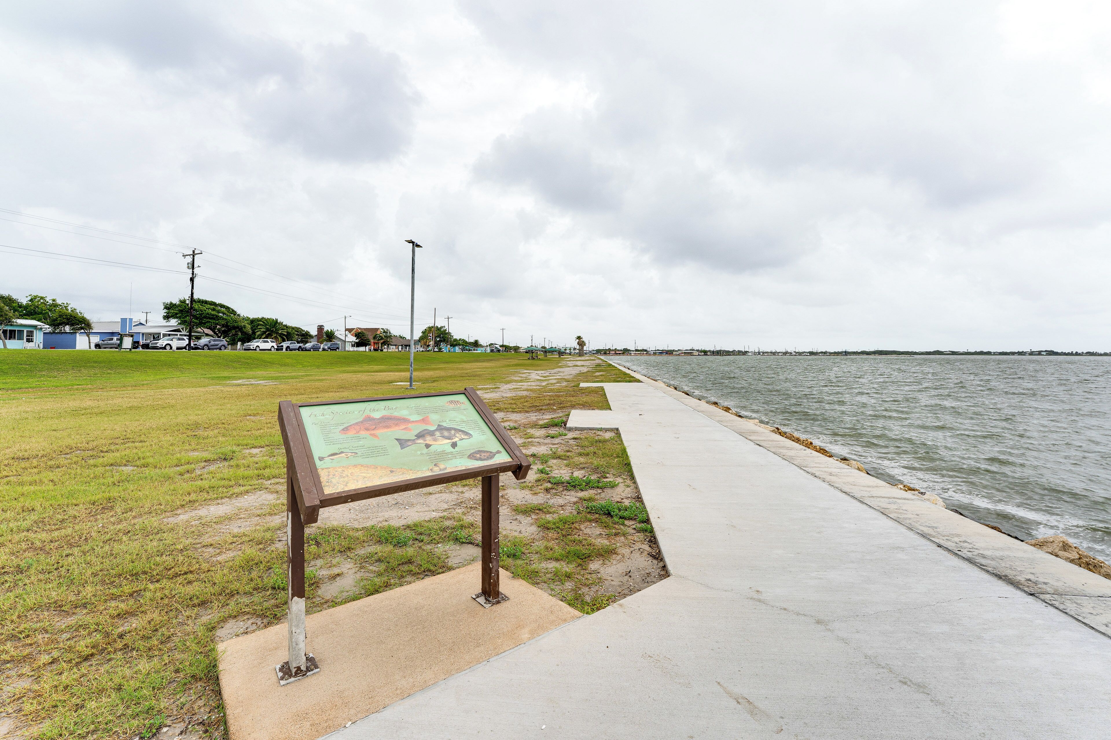 Une promenade en bord de mer, agrémentée d’un panneau d’information, d’une zone herbeuse et offrant une vue sur la mer.