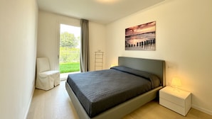 Een moderne slaapkamer met een bed, nachtkastje met lamp, raam met gordijnen en een schilderij aan de muur.