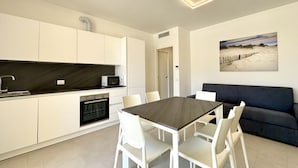 Une cuisine moderne avec des armoires blanches, un plan de travail foncé et un four encastré. On y trouve une table à manger avec des chaises blanches et un canapé foncé dans l’espace salon.