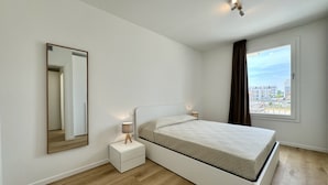 Une chambre moderne avec un lit simple, une table de chevet, un miroir et une fenêtre donnant sur des immeubles.