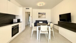 Cozinha moderna com armários brancos, forno embutido e micro-ondas. Há uma área de jantar com mesa e cadeiras, e uma televisão de tela plana fixada na parede.