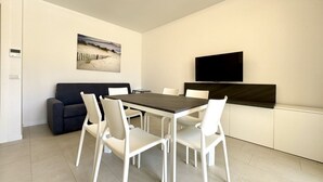 Uma sala de jantar moderna com uma mesa retangular e seis cadeiras brancas, um sofá escuro, uma TV de tela plana montada e uma obra de arte com tema de praia na parede.