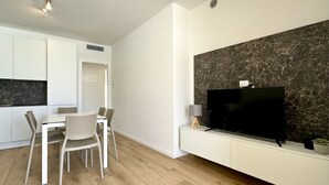 Un saló modern amb un televisor de pantalla plana fixat a una paret de marbre, una zona de menjador amb taula i cadires blanques, i una cuina amb armaris blancs i un taulell negre.