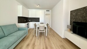 Un espace de vie moderne avec un canapé de couleur turquoise, un coin repas avec une table et des chaises blanches, une cuisine avec des meubles blancs et un grand téléviseur à écran plat posé sur un meuble blanc.