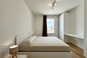 Ein kleines, modernes Schlafzimmer mit Bett, Nachttischlampe und Blick auf die Stadt durch ein Fenster.