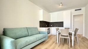 Een moderne woonkamer met een smaragdgroene bank, een eettafel met stoelen en een keuken met witte kasten en een zwarte achterwand.