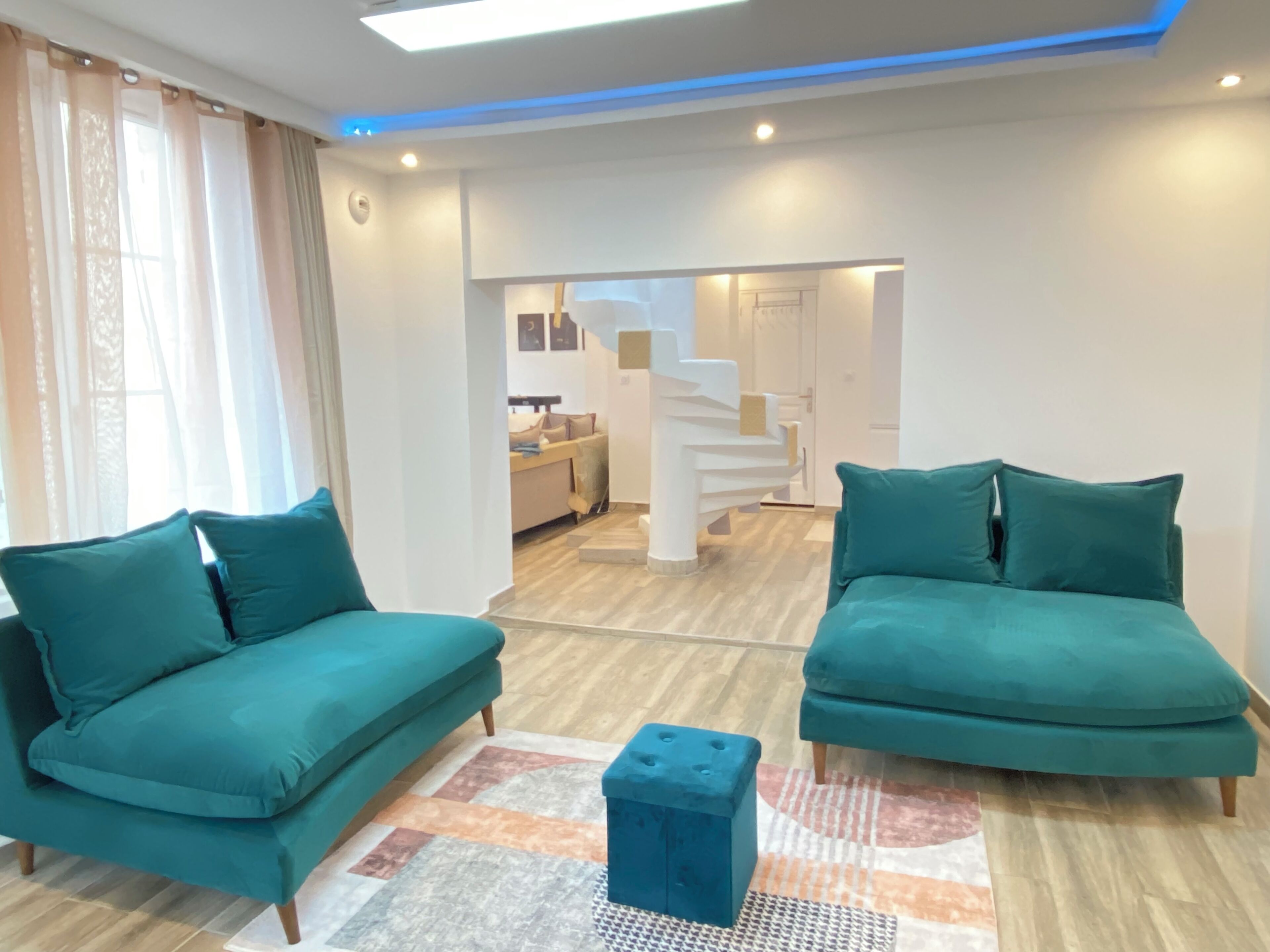 Ein modernes Wohnzimmer mit türkisfarbenen Sofas, einem zentralen Couchtisch und einer Treppe, die in eine obere Ebene führt.