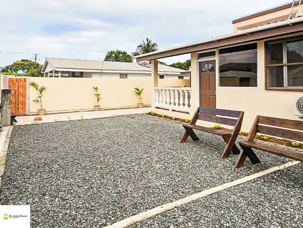 Une maison avec une allée en gravier, un banc en bois et un petit jardin agrémenté de plantes en pot.
