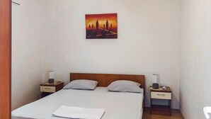 Une chambre d’hôtel avec un lit, deux tables de chevet, une commode, un tableau au mur et une chaise blanche.