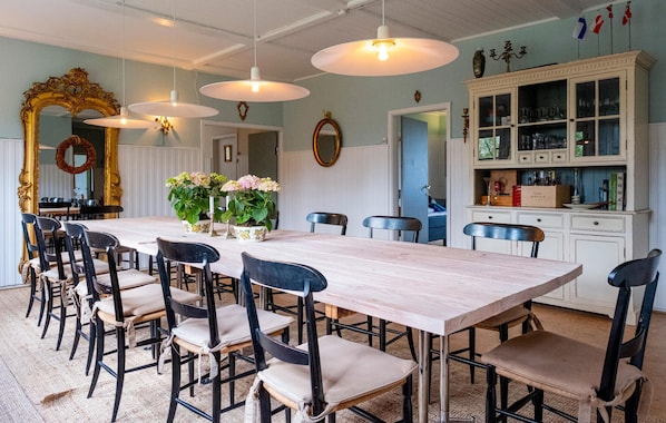 A dining area with a long table, multiple chairs, hanging pendant lights, and a cabinet with glass doors.