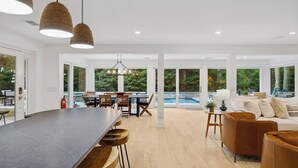 Un salón amplio con una gran mesa de madera oscura, sillas de cuero marrón, un sofá blanco y un reposapiés de cuero marrón. Hay un comedor con una mesa y sillas blancas, y una vista de una piscina a través de amplios ventanales.