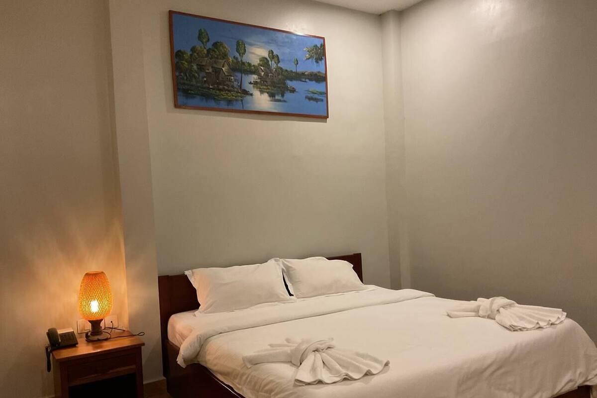 A neatly made bed with white linens and towels, a bedside lamp, and a painting of a lakeside scene on the wall.