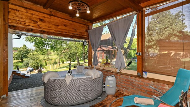 A cozy room with a wooden ceiling, a large window with curtains, a round bed, a blue chair, and a view of a green landscape.