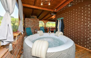 A hot tub in a rustic cabin with a brick wall, wooden ceiling beams, and a table with a bottle and glasses.